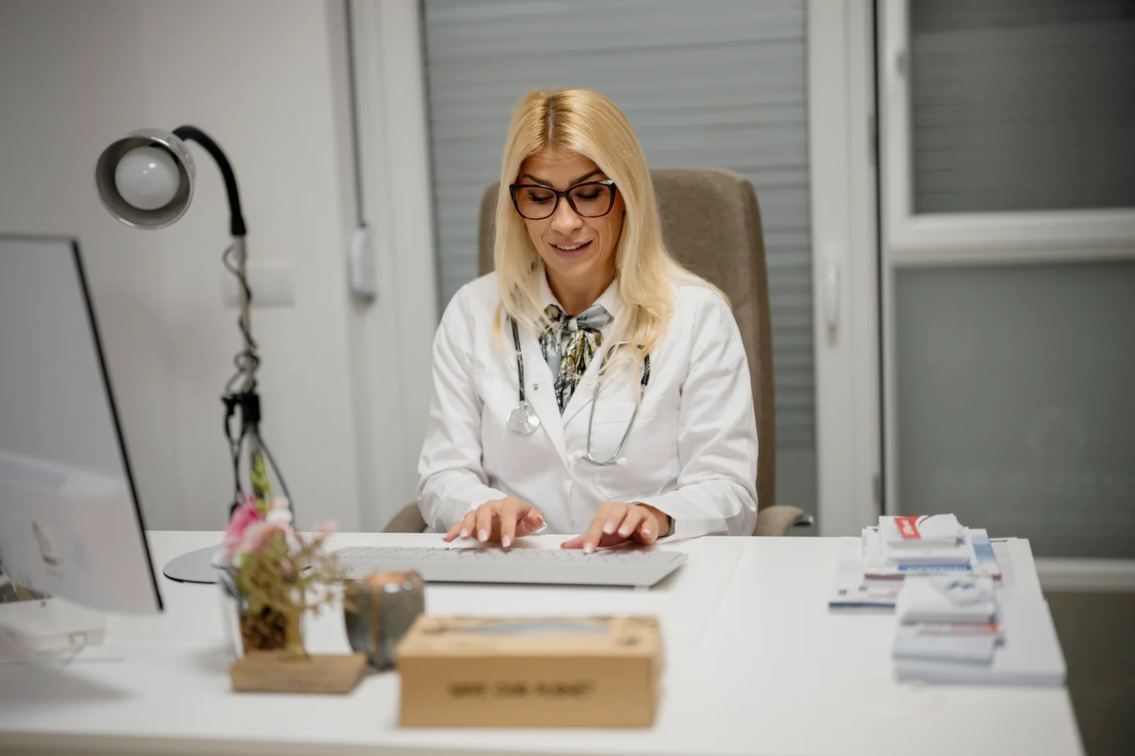 Person in a white coat and stethoscope typing at a desk